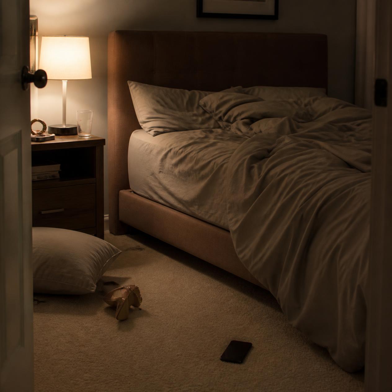 View into a bedroom with an unmade bed, a fallen pillow, a shoe on the floor and a phone near the bed.
