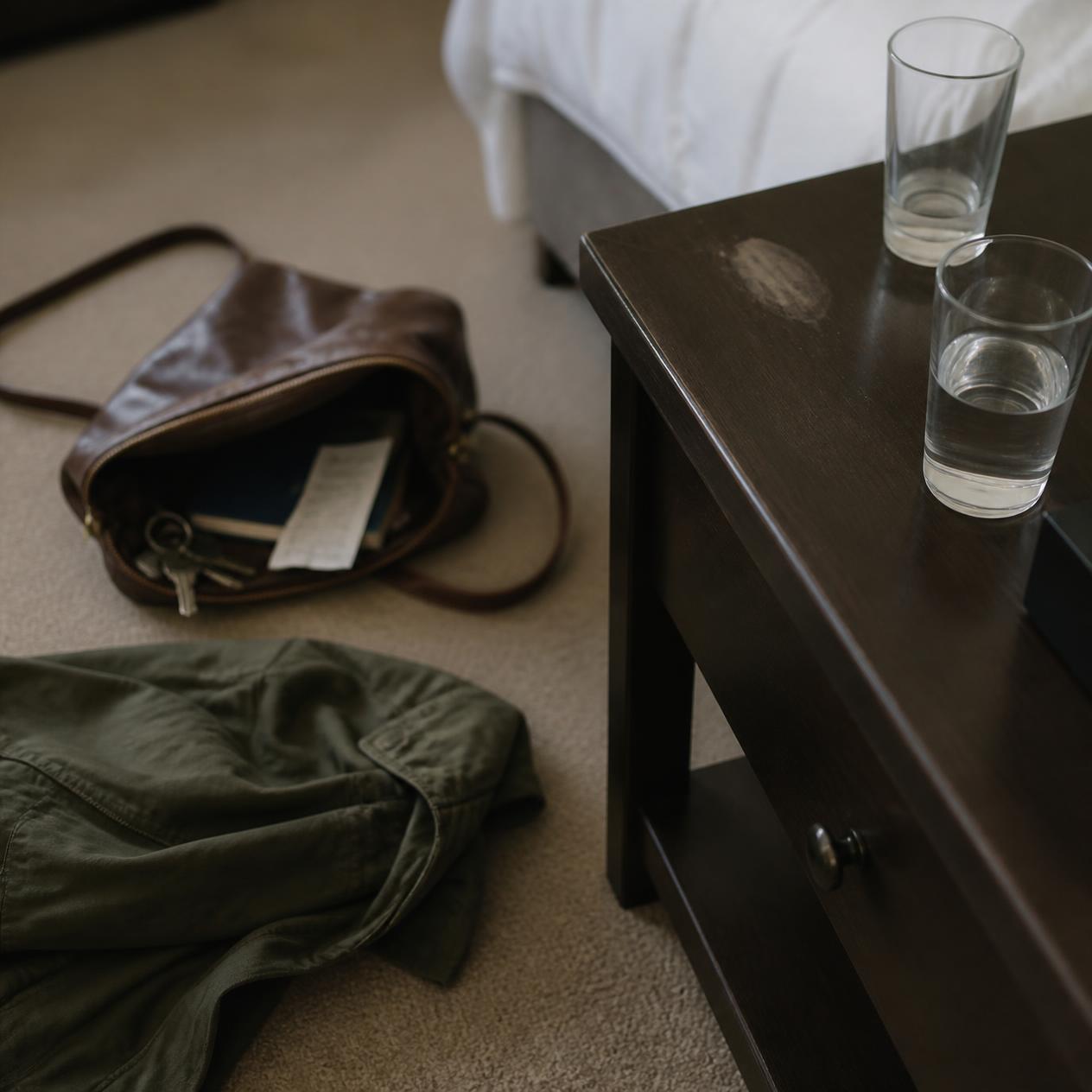 Close-up of a bedside table with two glasses, with an open bag and clothing on the floor nearby.